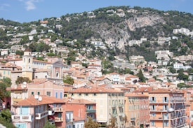 A picturesque hillside town with colorful buildings topped by terracotta roofs. The town is nestled against a lush green mountain, dotted with scattered white houses and abundant trees. The architecture reflects a Mediterranean style with pastel shades and elegant designs.
