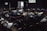 A dimly lit room filled with several people lying on the floor, appearing to be resting or injured. The scene has a somber atmosphere with light streaming in through a window and an open door, illuminating parts of the room.