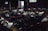 A dimly lit room filled with several people lying on the floor, appearing to be resting or injured. The scene has a somber atmosphere with light streaming in through a window and an open door, illuminating parts of the room.