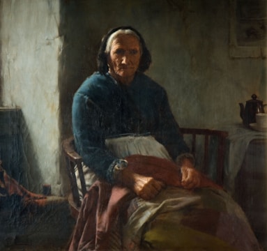 An elderly woman is seated in a dimly lit, rustic room. She wears a dark shawl and an apron while sitting in a wooden chair, possibly knitting or holding something in her hands. The room has a rough, worn appearance, with what looks to be a teapot and cup on a small table to the right side. The light appears to be coming from a window or an opening not fully visible on the left.