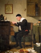 An elderly man with glasses and a black cap leans over an open desk drawer in a dimly lit room. He is surrounded by scattered papers and a ledger, suggesting an activity of accounting or scrutiny. The walls display a hanging map and a large key, indicating an interest in geography or possessions.