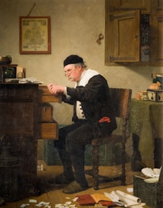 An elderly man with glasses and a black cap leans over an open desk drawer in a dimly lit room. He is surrounded by scattered papers and a ledger, suggesting an activity of accounting or scrutiny. The walls display a hanging map and a large key, indicating an interest in geography or possessions.