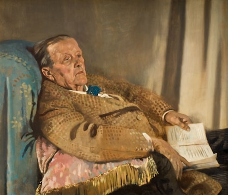 An older person is reclining in a chair with a patterned blanket. Their expression is contemplative as they hold an open book. The setting suggests a quiet, domestic scene with muted lighting.
