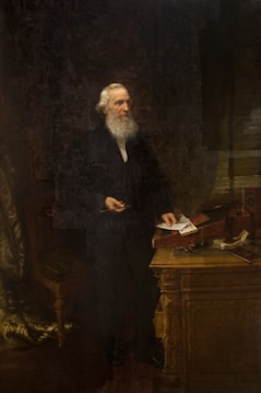 An elderly man with a long white beard stands beside a wooden desk, holding papers in one hand. The desk has various items on top, including a book and a decorative object. The man is wearing a dark suit, and the background suggests a dimly lit room with curtains drawn to one side.