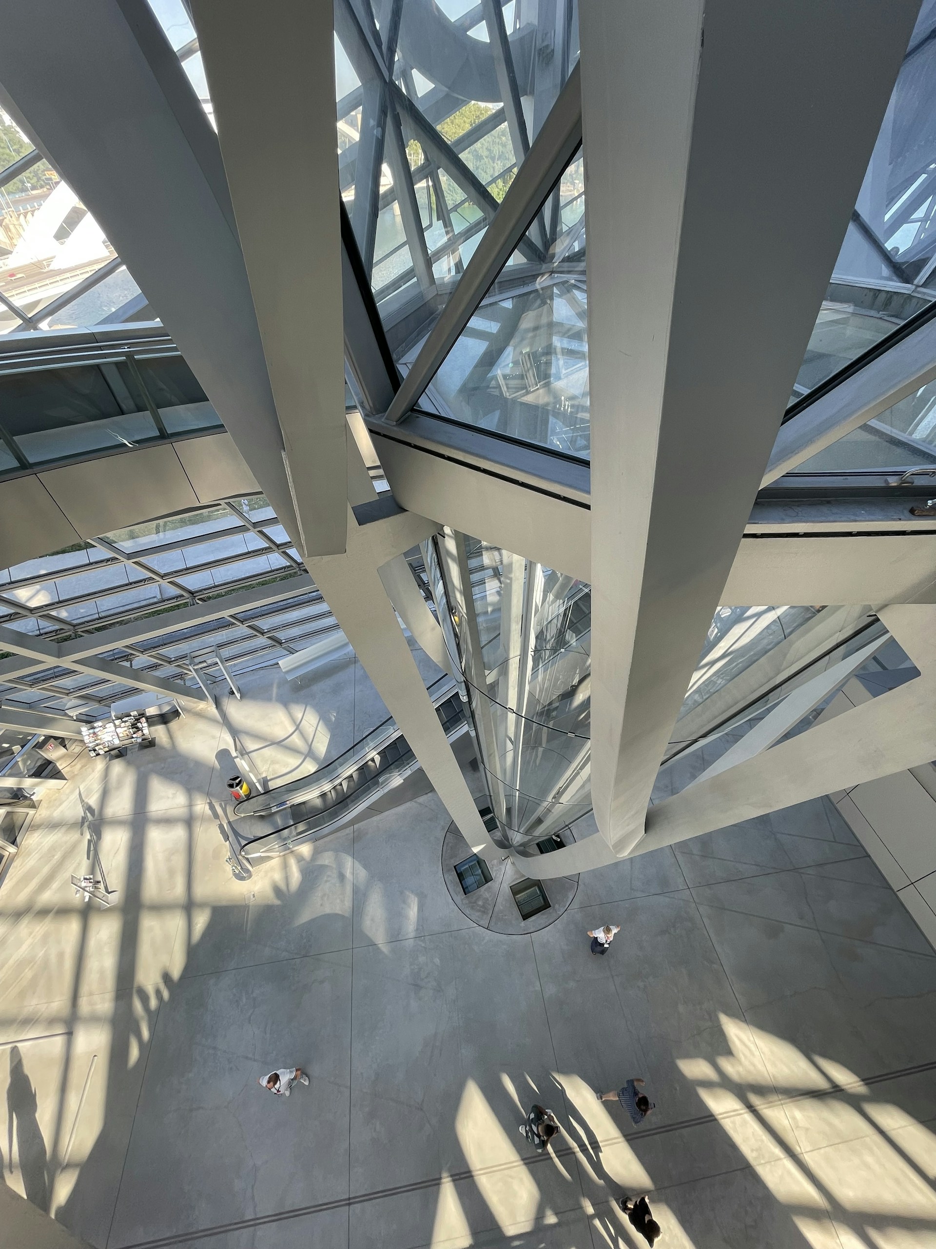 The image features the interior of a modern architectural structure with a view from above showing intersecting glass and metal beams. Sunlight creates dynamic patterns of light and shadow on the concrete floor. People can be seen walking, giving a sense of scale to the impressive design.