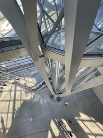 The image features the interior of a modern architectural structure with a view from above showing intersecting glass and metal beams. Sunlight creates dynamic patterns of light and shadow on the concrete floor. People can be seen walking, giving a sense of scale to the impressive design.