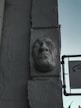 A sculpted face is embedded into the side of a weathered, textured wall. The face appears to be made of stone or plaster with a stern expression and closed eyes. To the right, there is a modern sign with a graphic depiction of a face. The overall lighting is subdued, with a slightly eerie atmosphere.