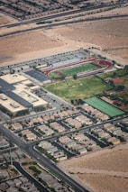 Aerial view of a suburban area featuring a school or sports complex. The facility includes a football field with a surrounding track, tennis courts, and several buildings. The surrounding residential area consists of organized rows of houses bordered by roads and desert terrain.