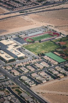 Aerial view of a suburban area featuring a school or sports complex. The facility includes a football field with a surrounding track, tennis courts, and several buildings. The surrounding residential area consists of organized rows of houses bordered by roads and desert terrain.