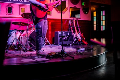 A musician is playing an acoustic guitar on a small stage with a drum set and several electric guitars in the background. The scene is lit with colorful, moody lighting, and the setting appears to be an intimate venue or music bar.