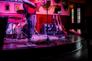 A musician is playing an acoustic guitar on a small stage with a drum set and several electric guitars in the background. The scene is lit with colorful, moody lighting, and the setting appears to be an intimate venue or music bar.