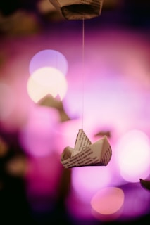 Paper boats made from printed text hang suspended by strings against a backdrop of vibrant pink and purple bokeh lights, creating a dreamy and whimsical atmosphere.