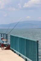A calm lakeside scene featuring a dock with fishing rods propped up against a teal railing. A young child wearing a blue cap and red shorts is seated on a white chair, seemingly focused on fishing. The expansive body of water extends towards distant mountains under a partly cloudy sky.