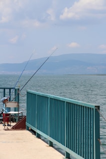 A calm lakeside scene featuring a dock with fishing rods propped up against a teal railing. A young child wearing a blue cap and red shorts is seated on a white chair, seemingly focused on fishing. The expansive body of water extends towards distant mountains under a partly cloudy sky.