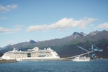 A large white cruise ship is docked near a shoreline with a small boat and a blue industrial crane nearby. Majestic mountains rise in the background under a clear sky scattered with clouds.