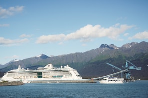 A large white cruise ship is docked near a shoreline with a small boat and a blue industrial crane nearby. Majestic mountains rise in the background under a clear sky scattered with clouds.