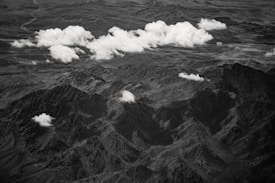 An aerial view of a rugged mountain range with scattered clouds casting shadows over the landscape. The terrain appears rocky and uneven, with a series of peaks and valleys stretching across the frame. The monochrome color palette adds a dramatic and timeless quality to the scene.