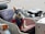 A small white dog is sitting in a sidecar attached to a vehicle. The dog is wearing a blue harness and protective goggles. The interior of the sidecar is upholstered and there is a spare tire attached to the back.