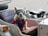 A small white dog is sitting in a sidecar attached to a vehicle. The dog is wearing a blue harness and protective goggles. The interior of the sidecar is upholstered and there is a spare tire attached to the back.