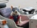 A small white dog is sitting in a sidecar attached to a vehicle. The dog is wearing a blue harness and protective goggles. The interior of the sidecar is upholstered and there is a spare tire attached to the back.