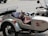 A small dog wearing goggles and a red vest is sitting in the sidecar of a motorcycle. The motorcycle is parked on a paved surface. The sidecar features a design with the words 'Russian Maiden' and a stylized graphic.