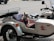 A small dog wearing goggles and a red vest is sitting in the sidecar of a motorcycle. The motorcycle is parked on a paved surface. The sidecar features a design with the words 'Russian Maiden' and a stylized graphic.