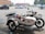 A white dog wearing a red vest is sitting in the sidecar of a motorcycle parked in a parking lot. The motorcycle has a retro design and is equipped with a spare tire mounted on the back of the sidecar. Several cars are parked in the background near some buildings and trees.