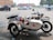 A white dog wearing a red vest is sitting in the sidecar of a motorcycle parked in a parking lot. The motorcycle has a retro design and is equipped with a spare tire mounted on the back of the sidecar. Several cars are parked in the background near some buildings and trees.