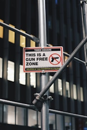 A sign indicating a 'Gun Free Zone' is mounted on a metal scaffolding structure. The sign is located in Times Square and includes a graphic of a handgun with a red circle and slash over it. The building in the background has dark vertical lines and illuminated windows.