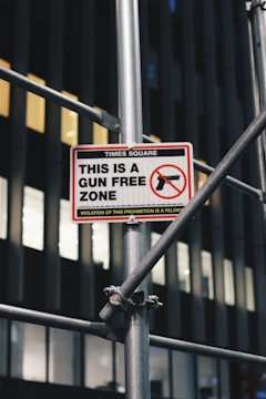A sign indicating a 'Gun Free Zone' is mounted on a metal scaffolding structure. The sign is located in Times Square and includes a graphic of a handgun with a red circle and slash over it. The building in the background has dark vertical lines and illuminated windows.