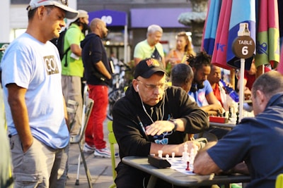 A group of people engaged in a chess game outdoors, with several chess boards set up on tables under colorful umbrellas. One man appears to be making a move while another watches intently. Spectators stand and sit around, creating a lively and focused atmosphere.