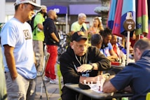 A group of people engaged in a chess game outdoors, with several chess boards set up on tables under colorful umbrellas. One man appears to be making a move while another watches intently. Spectators stand and sit around, creating a lively and focused atmosphere.