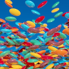 A vibrant collection of multi-colored round candies appears to be floating against a bright blue background. The candies include a variety of colors like yellow, red, blue, orange, and green, creating a dynamic and playful visual effect.