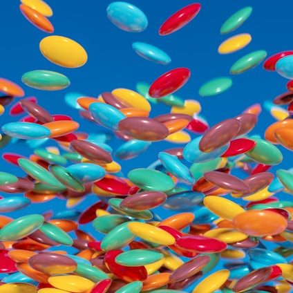 A vibrant collection of multi-colored round candies appears to be floating against a bright blue background. The candies include a variety of colors like yellow, red, blue, orange, and green, creating a dynamic and playful visual effect.