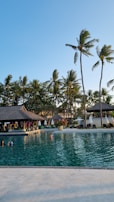 A serene swimming pool with clear water surrounded by tropical palm trees. There is a thatched-roof pavilion with sun loungers and umbrellas lining the poolside. Several people are enjoying the pool under the bright, clear blue sky.