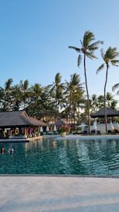 A serene swimming pool with clear water surrounded by tropical palm trees. There is a thatched-roof pavilion with sun loungers and umbrellas lining the poolside. Several people are enjoying the pool under the bright, clear blue sky.