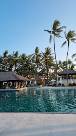 A serene swimming pool with clear water surrounded by tropical palm trees. There is a thatched-roof pavilion with sun loungers and umbrellas lining the poolside. Several people are enjoying the pool under the bright, clear blue sky.