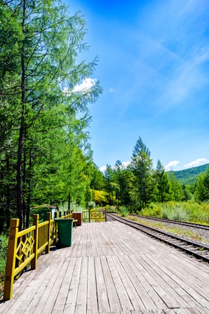 A wooden platform stretches alongside a railway track, surrounded by lush green trees under a bright blue sky. The scene is likely in a rural or forested area with no visible people. There are yellow railings and a green trash can on the platform, with hills visible in the distance.