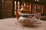 A yellow ceramic teapot with a curved spout rests on a sandy-colored surface, accompanied by a small, ornately painted ceramic container with a blue and white pattern. A clear glass sits nearby, with remnants of liquid visible at the bottom. The background consists of a blurred wooden structure that provides a warm, rustic ambiance.