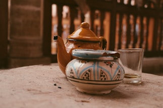 A yellow ceramic teapot with a curved spout rests on a sandy-colored surface, accompanied by a small, ornately painted ceramic container with a blue and white pattern. A clear glass sits nearby, with remnants of liquid visible at the bottom. The background consists of a blurred wooden structure that provides a warm, rustic ambiance.
