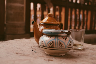 A yellow ceramic teapot with a curved spout rests on a sandy-colored surface, accompanied by a small, ornately painted ceramic container with a blue and white pattern. A clear glass sits nearby, with remnants of liquid visible at the bottom. The background consists of a blurred wooden structure that provides a warm, rustic ambiance.