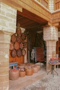 A rustic market stall showcases an array of traditional handicrafts, including decorative woven baskets and colorful textiles. The scene is set under a wooden canopy with stone pillars, and the ground is cobbled. Various handcrafted items are neatly arranged, featuring intricate patterns and vibrant colors.