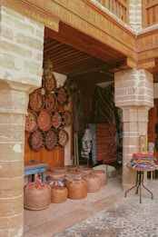A rustic market stall showcases an array of traditional handicrafts, including decorative woven baskets and colorful textiles. The scene is set under a wooden canopy with stone pillars, and the ground is cobbled. Various handcrafted items are neatly arranged, featuring intricate patterns and vibrant colors.
