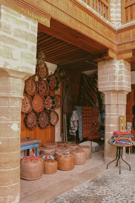 A rustic market stall showcases an array of traditional handicrafts, including decorative woven baskets and colorful textiles. The scene is set under a wooden canopy with stone pillars, and the ground is cobbled. Various handcrafted items are neatly arranged, featuring intricate patterns and vibrant colors.