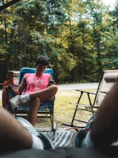 A person is sitting in a blue camping chair, wearing a pink sleeveless shirt and holding a phone. The scene is set outdoors with lush green trees in the background, accompanied by a calming atmosphere. The legs of another person are visible in the foreground, suggesting a relaxed setting. Adjacent to the seated individual, there is a black folding chair and the edge of a beige lounge chair, all situated on a patterned rug.