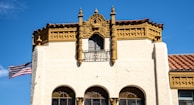 A historic building with ornate architectural details, including decorative carvings and columns, with a beige facade and a red-tile roof. An American flag is waving prominently on the left side of the structure against a clear blue sky.