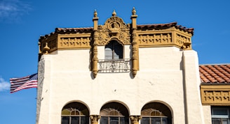 A historic building with ornate architectural details, including decorative carvings and columns, with a beige facade and a red-tile roof. An American flag is waving prominently on the left side of the structure against a clear blue sky.