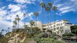 A luxurious hillside building with cream-colored walls is surrounded by lush greenery and tall palm trees. The sky above is bright blue with scattered white clouds, and people are walking along a landscaped pathway leading up to the building.