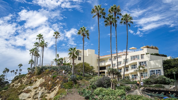 A luxurious hillside building with cream-colored walls is surrounded by lush greenery and tall palm trees. The sky above is bright blue with scattered white clouds, and people are walking along a landscaped pathway leading up to the building.