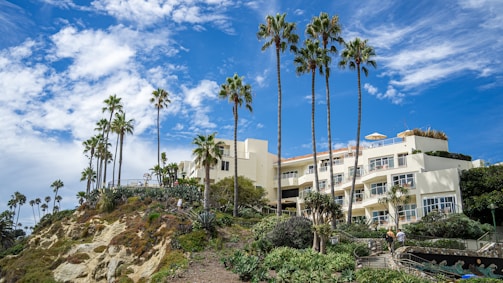 A luxurious hillside building with cream-colored walls is surrounded by lush greenery and tall palm trees. The sky above is bright blue with scattered white clouds, and people are walking along a landscaped pathway leading up to the building.
